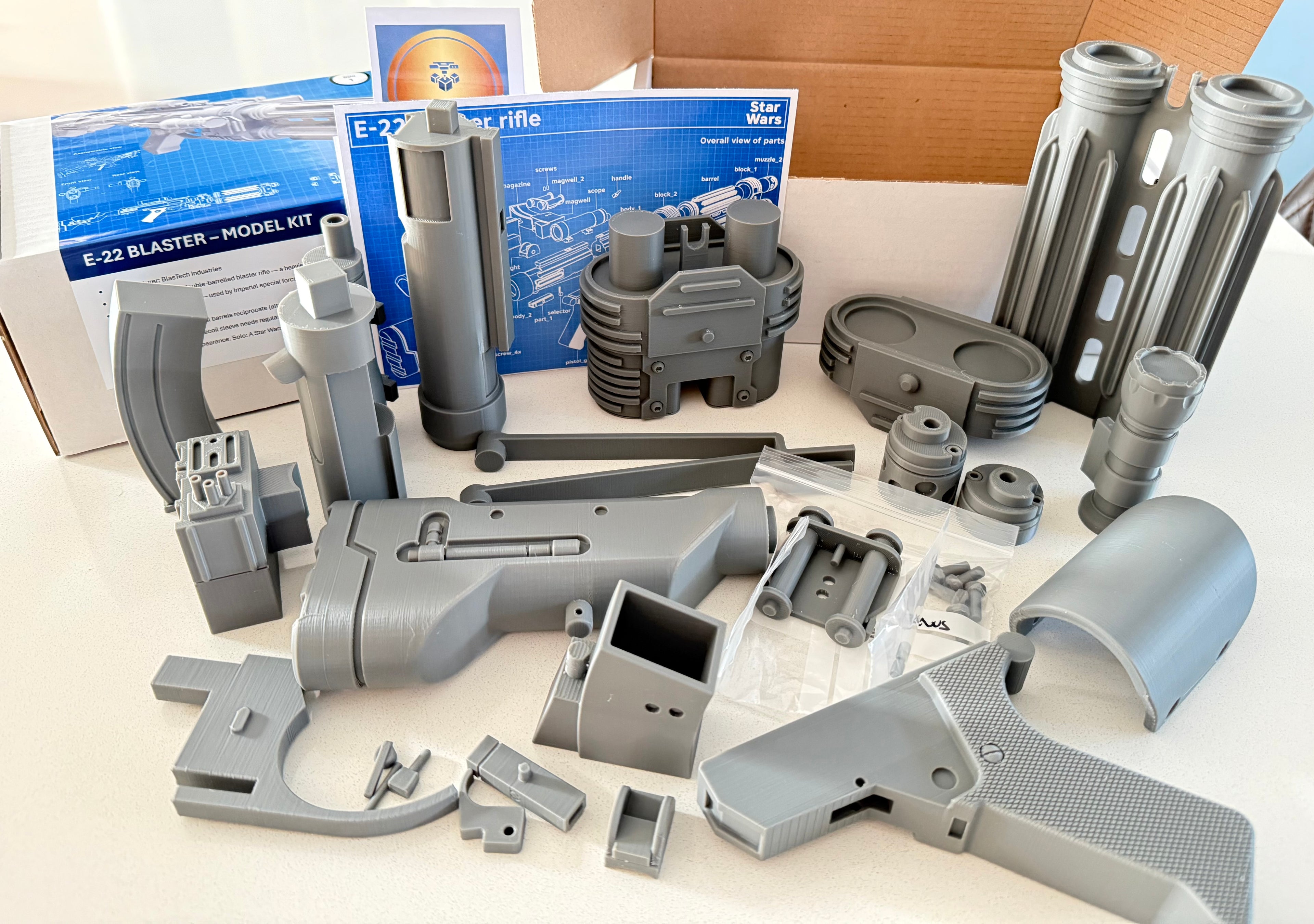 A 3D-printed replica of an E-22 Blaster from the E-22 Rifle model kit by Star Citizen, shown in its entirety with various disassembled parts and accessories spread out on a white surface, alongside its blue and white retail packaging.