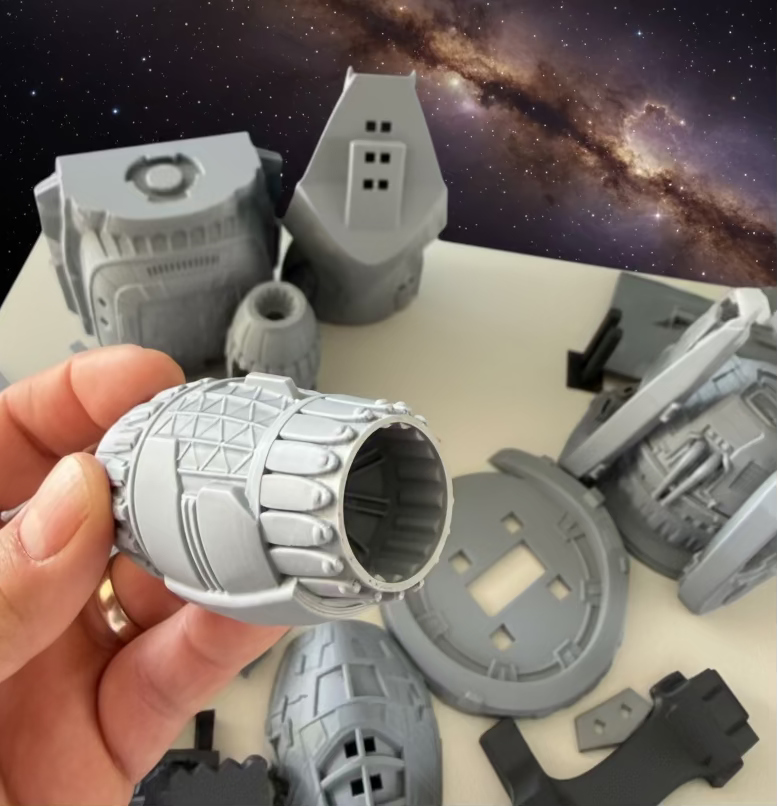 A hand holding a gray 3D-printed sci-fi cylindrical component, with other unpainted model parts scattered on a white surface against a blue background.