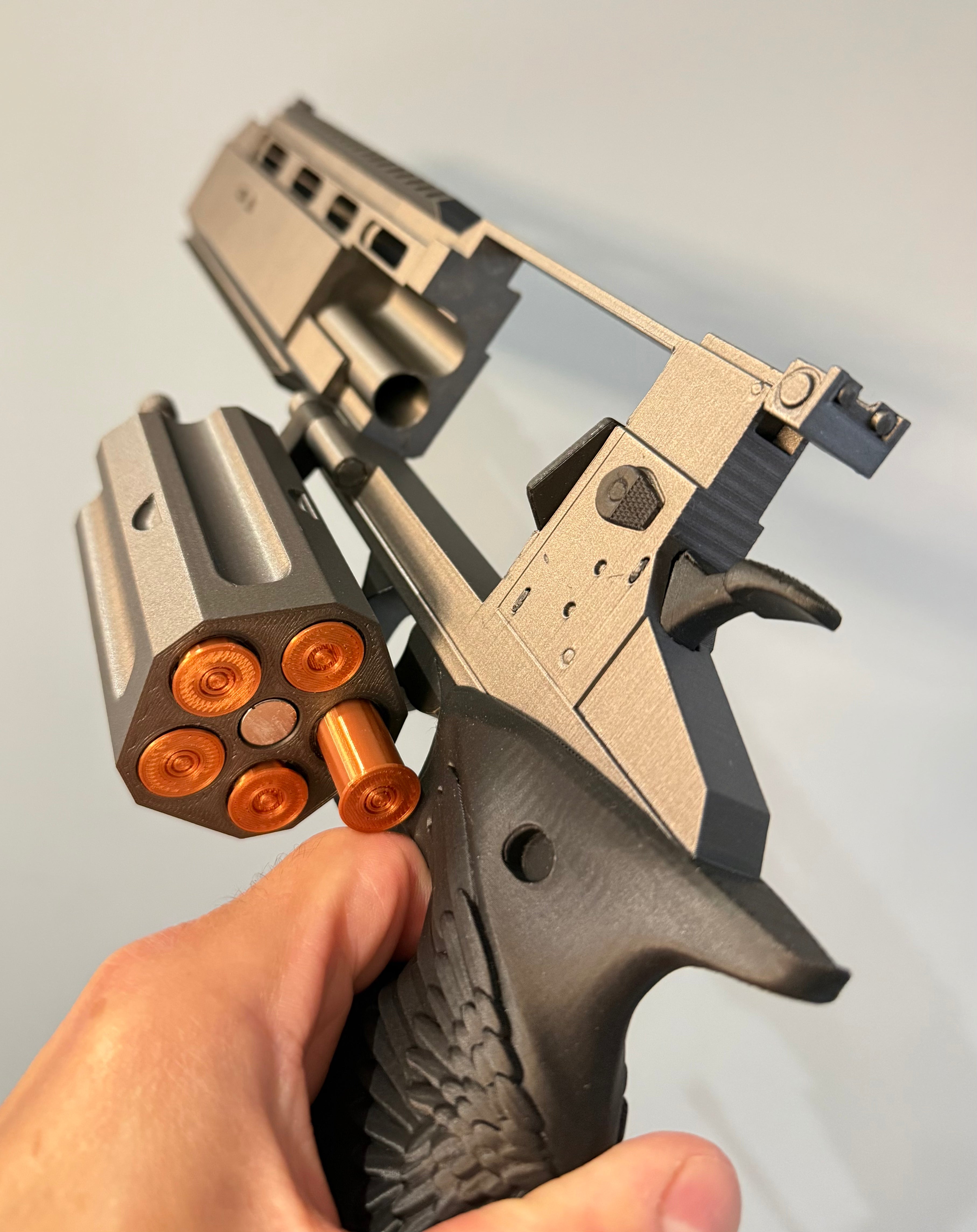 A close-up of a hand holding a metallic silver and black revolver-style toy oLeon's Magnum Revolver – Resident Evil Requiem Prop Kit with metallic copper bullet casings visible in the cylinder, against a plain light background.