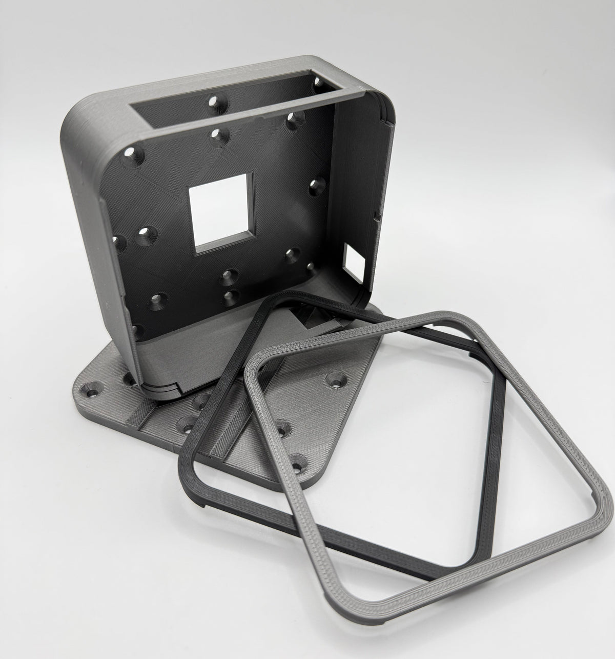 Hive Thermostat in dark grey metallic mounting bracket with a square cut-out, shown alongside two matching square metal frame pieces on a light grey studio background.